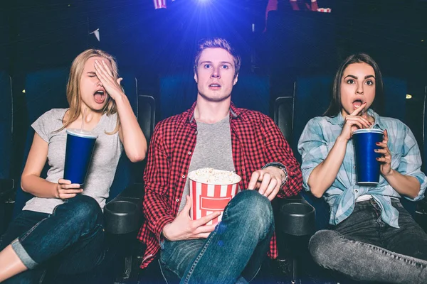 Youn man and women are sitting in chairs and watching movie. They are surprised and confused. Blonde girl put her hand on face. Brunette girl stopped drinking cola. Guy is holding a basket of popcorn. — Stock Photo, Image