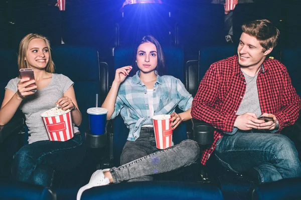 Very good young friends are sitting together in cinema. Blonde gir and boy are looking at each other and smiling. She is holding phone in hands. Brunette is looking straight and eating popcorn. — Stock Photo, Image