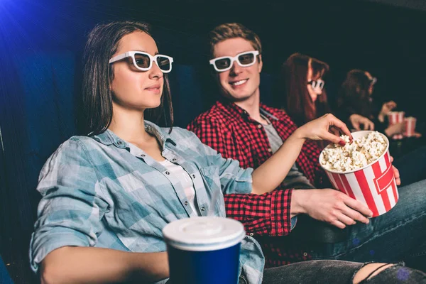 A picture of company sitting together in one row in cinema hall. Girl is watching movie in glasses and reaching the basket with popcorn that her boyfriend has. He is looking at her and smiling.