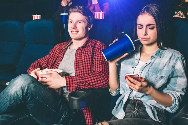 Abgelenktes Mädchen schaut am Telefon zu. Sie langweilt sich beim Ansehen von Filmen. Außerdem trinkt sie Cola aus einem großen blauen Becher. Kerl ist glücklich und guckt Film. — Stockfoto