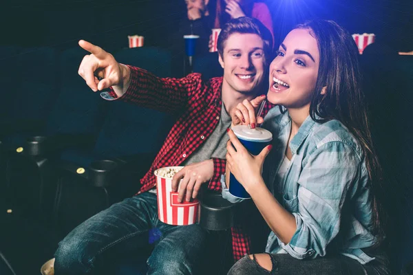 Amazing and cheerful girl is laughing. She is drinking coke from blue cup. Guy wants to grab her attention and pointing on camera. He is looking ar her. — Stock Photo, Image