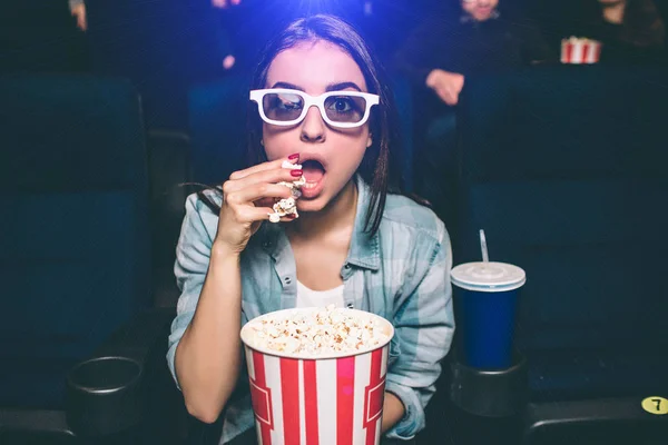 Удивительная девушка сидит очень близко и смотрит прямо. Она носит специальные 3D очки для просмотра фильмов. Девочка ест попкорн правой рукой. Кроме нее, есть еще чашка кокса. . — стоковое фото