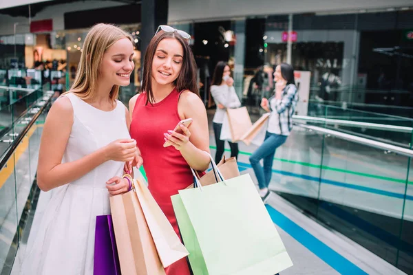 Attraktive und schöne Mädchen stehen mit Taschen zusammen. brünettes Mädchen hält Telefon und schaut es sich zusammen mit ihrem Freund an. Blondes Mädchen lächelt. ihre Freunde stehen hinter ihnen. — Stockfoto