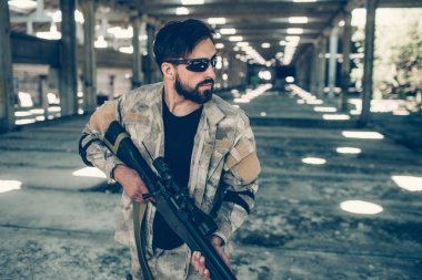 Ciddi ve yakışıklı bir adam gözlüklü hangarda duran ve paintball silahı elinde tutan. Düz ön bakıyor. Adam özel üniforma giyer.