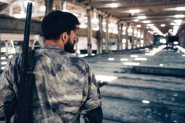 Bearded brunette is standing in hude and long hangar. He is standing back to camera. Guy is looking to the right. There is rifle hanging over his shoulder.