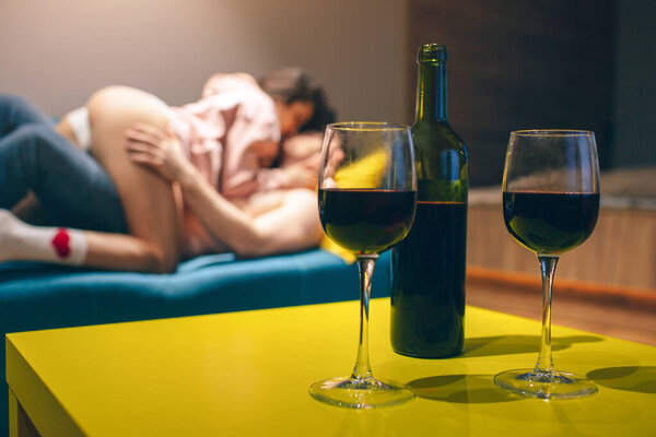 Young couple have intimacy in kitchen in night. Seductive sensual people in sex position on sofa. Wine bottle stand on table with glasses.