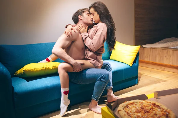 Mladý pár má v noci intimitu v kuchyni. Vášnivý muž bez trička drží ženu na nohou a líbá ji. Svůdný smyslný model v sexuální pozici těší. Pizza na stole. — Stock fotografie
