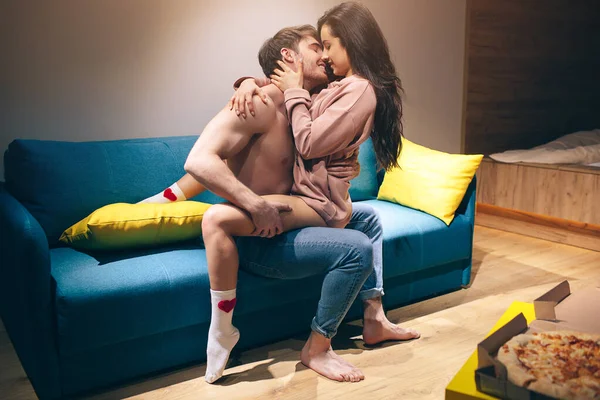 Unga par har intimitet i köket på natten. Passionerad tröjlös man sitter på soffan och kysser sensuell kvinna. Förförisk sexig modell sitter på hans ben och omfamnar killen. Pizza på bordet. — Stockfoto