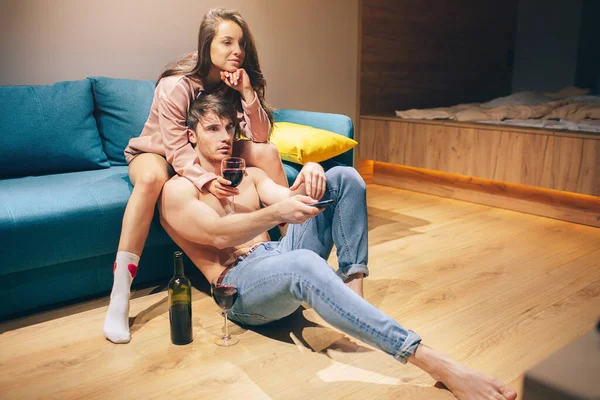 Молодая сексуальная пара после интимной жизни на кухне ночью. Мужчина без рубашки сидит на полу и смотрит вперед. Соблазнительная женщина на диване и обнимает его. Питьевое вино . — стоковое фото