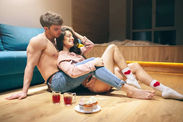 Młoda seksowna para po intymności w kuchni w nocy. Kobieta siedzi na męskich nogach i uśmiecha się. Facet bez koszuli patrzy na modelkę. Kubki herbaty i ciasta na podłodze. — Zdjęcie stockowe
