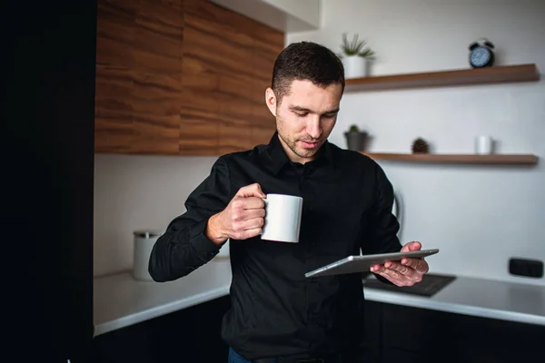 Un joven empresario trabaja en la cocina. Empresario estar solo, sostener la tableta y mirarlo. El hombre de negocios tiene un día productivo. Pausa para café o descanso . — Foto de Stock