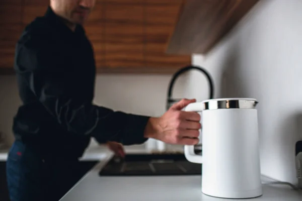 Jeune homme d'affaires en chemise noire stand dans la cuisine et tenir blanc bouilloire électrique intelligente. Il veut chauffer de l'eau. Tenez le gros doigt sur le bouton. Prêt à chauffer l'eau. Pause café . — Photo