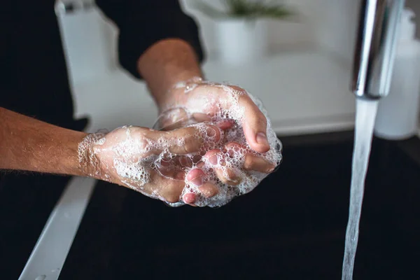 Feche de mãos de homem em espuma de sabão. Lavar os dedos e palmas das mãos e limpá-los. Água a sair da torneira para o lavatório na cozinha. Cuidados com as mãos e proteção. cara desgaste preto camisa . — Fotografia de Stock