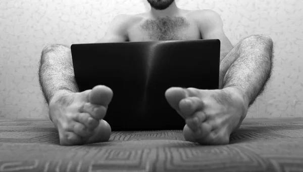 Imagen de hombre joven desnudo con las piernas peludas sentarse en el sofá y sostener el ordenador portátil entre los pies. Cúbrete con la pantalla del dispositivo. Vista baja. Cuadro en blanco y negro. Vista de corte . — Foto de Stock