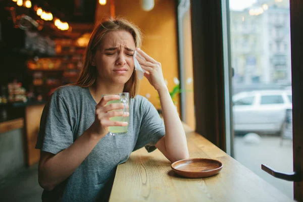 Μια νεαρή ξανθιά σε ένα καφέ. Έχει κρύωμα και μύτη που τρέχει, πονάει το κεφάλι της. Κρατάει ένα φλιτζάνι ζεστό τσάι. — Φωτογραφία Αρχείου