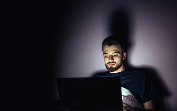 Young man lying on bed and look at laptop screen. Calm concentrated guy working overtime playing games or entertain himself. Night room. Isolated over dark background. — Stock Photo, Image