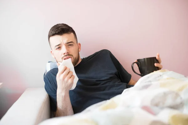 Young ill man with hanky. Guy lying on the sofa under covers. He holds napkin in one hand and black cup in other. Begins to sneeze. Isolated over light background.
