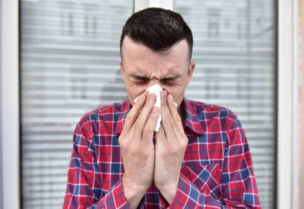 Молодой парень в рубашке чихает на белую салфетку. Заболела, подхватила простуду или коронавирус. Грипп и головная боль. Плохие симптомы. Встань снаружи в окошко . — стоковое фото