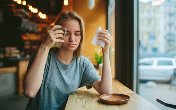 Young blonde in a cafe. She has a cold and a runny nose, her head hurts. She holds a cup of tea near her head — Stock Image Young blonde in a cafe. She has a cold and a runny nose, her head hurts. She holds a cup of tea near her head Stock Picture