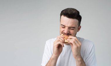 Young man isolated over background. Picture of guy eating burger and biting it. Tasty delisious fatning meal. Eating with pleasure. No guilty for eating bad food. Alone in studio. clipart
