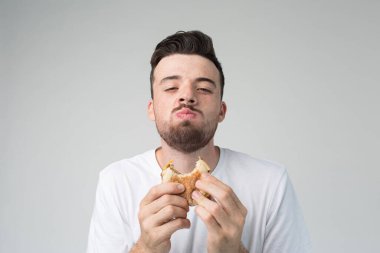 Genç adam arka planda izole edilmiş. Mutlu adam kameraya bakıp hamburger çiğniyor. Kötü şişmanlatan yemekleri zevkle yemek. Fast food ellerinde..