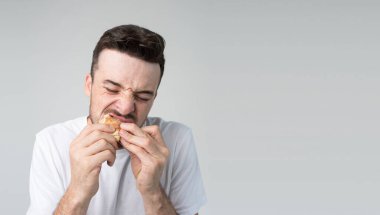 Genç adam arka planda izole edilmiş. Stüdyoda ısırıp çiğneyen lezzetli bir burger. Zevk duydum. Lezzetli yemek. Sağlıksız, şişman, kötü yemek..