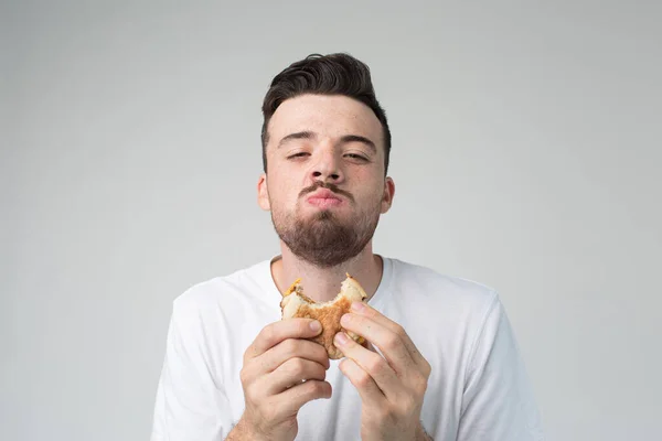 Mladý muž izolovaný v pozadí. Šťastný spokojený chlap se dívá na kameru a žvýká burger. Jíst špatné výkrm jídlo s radostí. Rychlé občerstvení v rukou. — Stock fotografie