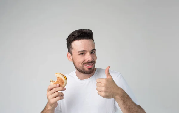 Young man isolated over background. Cheerful positive guy hold piece of bunger in hand and show big thumb up. Eating fastfood but like it. Delisious tasty meal. Alone on background. — Stock Photo, Image
