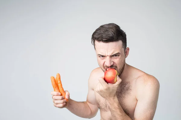 그는 이렇게 말합니다. 셔츠를 입지 않은 수컷 채식주의자가 사과를 깨물며 카메라를 보는 표정. 오렌지 당근을 손에 쥐고. 잔인 한 사람의 포즈. — 스톡 사진