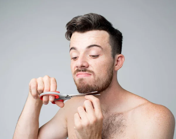 Jovem isolado sobre o fundo. Retrato de um tipo a tentar cortar a barba com uma tesoura. Auto-barbeiro. Olhe para baixo e faça isso com precisão e gentileza . — Fotografia de Stock