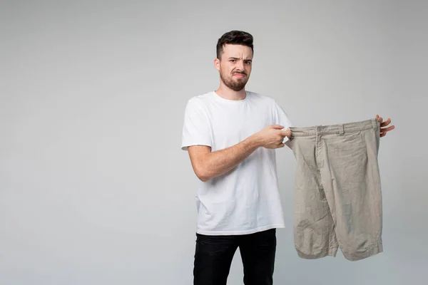 Junger Mann isoliert über Hintergrund. Kerl im lässigen Look hält große Hosen in den Händen. Gesichtsausdruck des Abscheus. Fatale Hose von jemandem. — Stockfoto