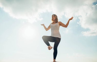 Genç kadın dışarıda yoga yapıyor. Sarışın kız bir bacağının üstünde duruyor, diğeri de dizlerinin üstüne çökmüş. Ellerinizi dirseklerinize kadar bükün ve yukarı dönün. Arkaplanda gökyüzü ve bulutlar.