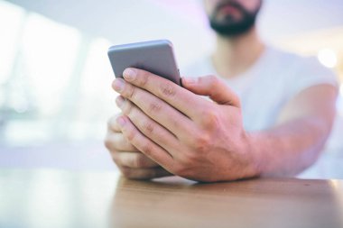 Sakallı genç bir adamın yan görünüşü, üstünkörü giyinmiş, kafenin yuvarlak ahşap masasında oturuyor ve elinde akıllı telefon var..
