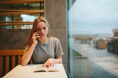 Sarışın kitap okuyor ve bir lokantada ya da restoranda telefonla konuşuyor.