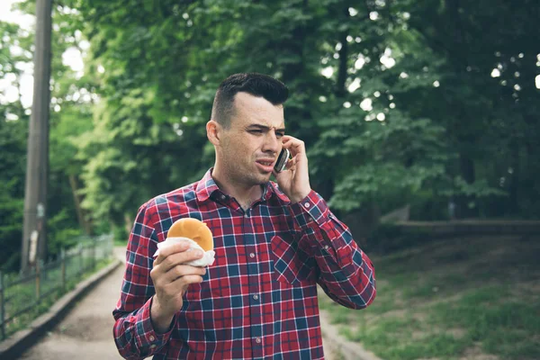 Un joven guapo comiendo sándwich a domicilio. Está sosteniendo un teléfono. — Foto de Stock
