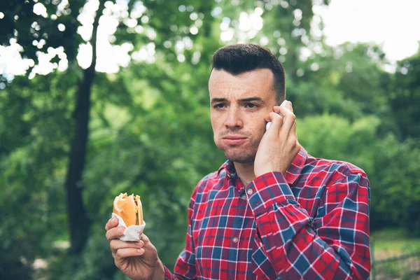 Knappe jongeman die een broodje autdoor eet. Hij heeft een telefoon vast. — Stockfoto