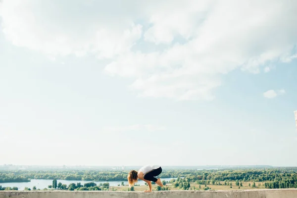 Ung kvinna utövar yoga utanför. Blond flicka gör kran pose eller bakasana på parapet på vacker utsikt. Hon klädde sig i svarta leggings och vit t-shirt. Träd flod och himmel på bakgrunden. — Stockfoto