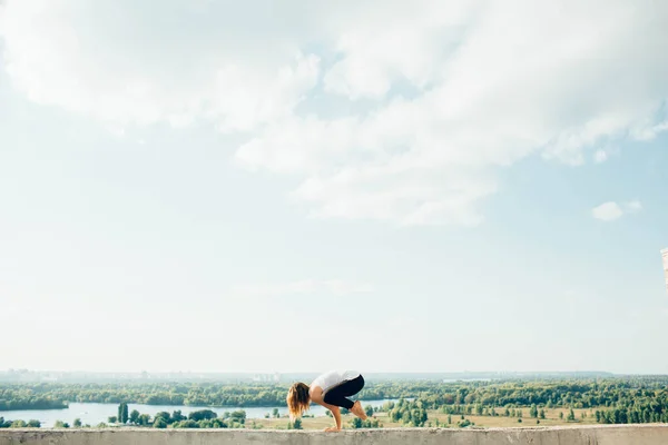 Junge Frau praktiziert Yoga draußen. Blonde Mädchen tun Kran Pose oder Bakasana auf Brüstung auf schöne Aussicht. Sie trug schwarze Leggings und ein weißes T-Shirt. Bäume Fluss und Himmel im Hintergrund. — Stockfoto