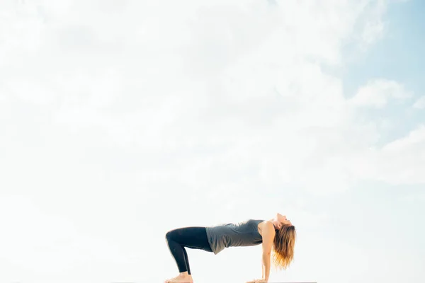 Junge Frau praktiziert Yoga draußen. Blonde Mädchen posieren auf dem Kopf. Purvottanasana Yoga-Haltung. Bekleidet war sie mit schwarzen Leggings und grauem T-Shirt. Himmel und Wolke im Hintergrund. — Stockfoto