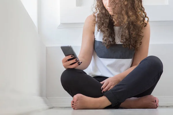 在白色的房间里,一个卷发年轻女子坐在地板上的剪影.拿起智能手机并使用它。现代技术和电话设备. — 图库照片