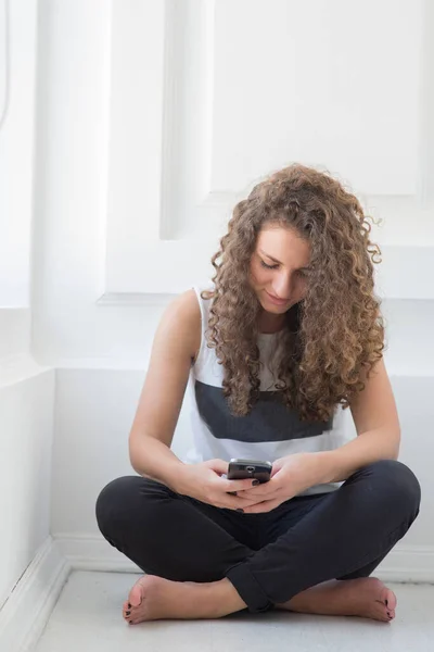 年轻的卷发女子坐在地板上使用智能手机的垂直图片。数字技术和现代装置。一个人坐在白色房间里. — 图库照片