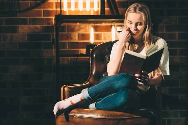 Menina em um quarto escuro acolhedor lendo um livro com óculos. Luz de lâmpada loira e quente — Fotografia de Stock