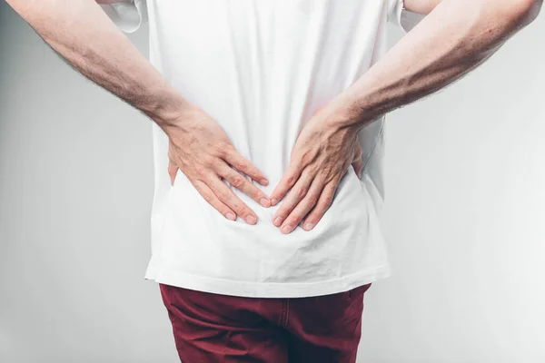 Caucasian man in white T-shirt and burgundy pants. His hands holding on lower back. Cut view. Concept. Loins hurts. Isolated over light background. — Stock Photo, Image