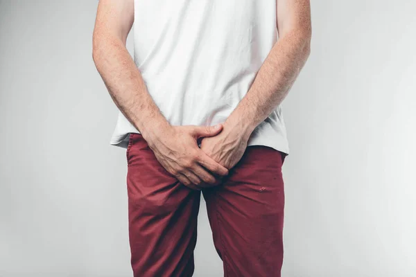 Caucasian man in white T-shirt and burgundy pants. Holds his hands on groin. Cut view. Concept. Isolated over light background. — Stock Photo, Image