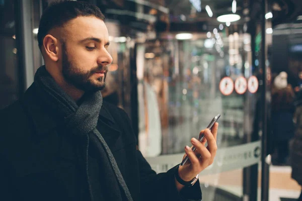 Vista lateral del joven barbudo, vestido con ropa informal, que está de pie en la calle y utilizando un teléfono inteligente — Foto de Stock