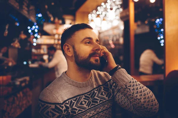 Side view of young bearded man,dressed incasual wear,sitting at round wooden table in cafe with modern interior and is holding smartphone. — Stock Photo, Image