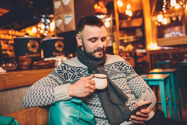 stock image Side view of young bearded man,dressed incasual wear,sitting at round wooden table in cafe with modern interior and is holding smartphone.