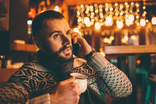 Vue latérale du jeune homme barbu, habillé vêtements décontractés, assis à une table ronde en bois dans un café avec un intérieur moderne et tient smartphone . — Photo