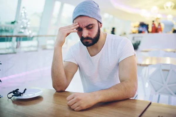 Tired young man has headache. Sit in cafe and hold hand on head. Hipster caught cold or pain in head. Sit inside restaurant or odern cafe. — Stock Photo, Image