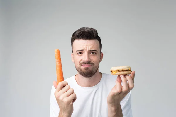 O rapaz come uma cenoura e um hambúrguer. O homem faz a escolha entre comida rápida e saudável. Saboroso ou útil O dilema de escolher um estilo de vida diferente — Fotografia de Stock
